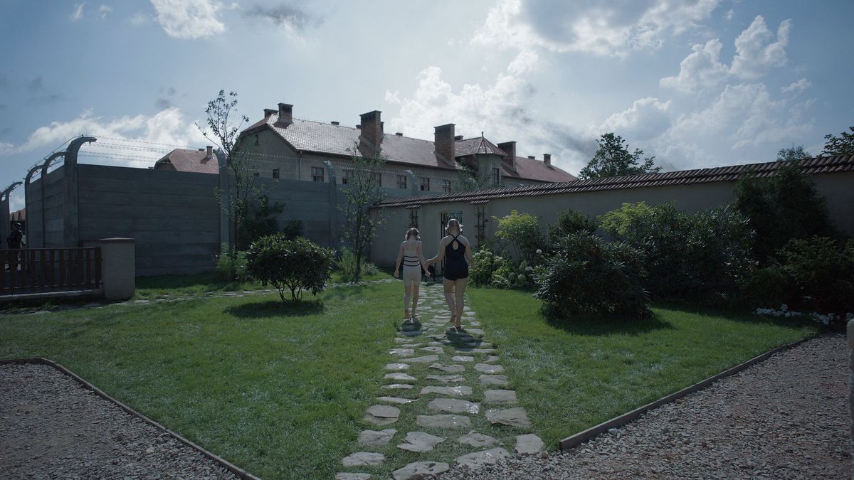 Two girls walk down a path in a walled garden with the boarding houses of Auschwitz looming above them in The Zone of Interest