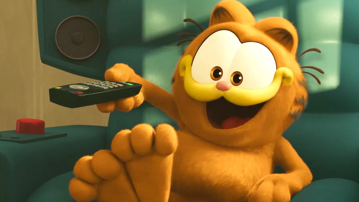 Garfield smiles while changing the station with the TV remote while sitting in The Garfield Movie