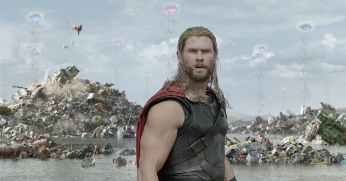 Thor looks around puzzled in what looks like a giant trash heap. In the sky behind him portals can be seen, with trash falling from them to the ground. 