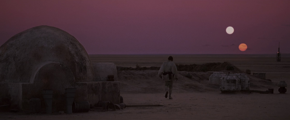 Luke Skywalker walks across his sandy yard towards the horizon, two suns setting in the sky. 
