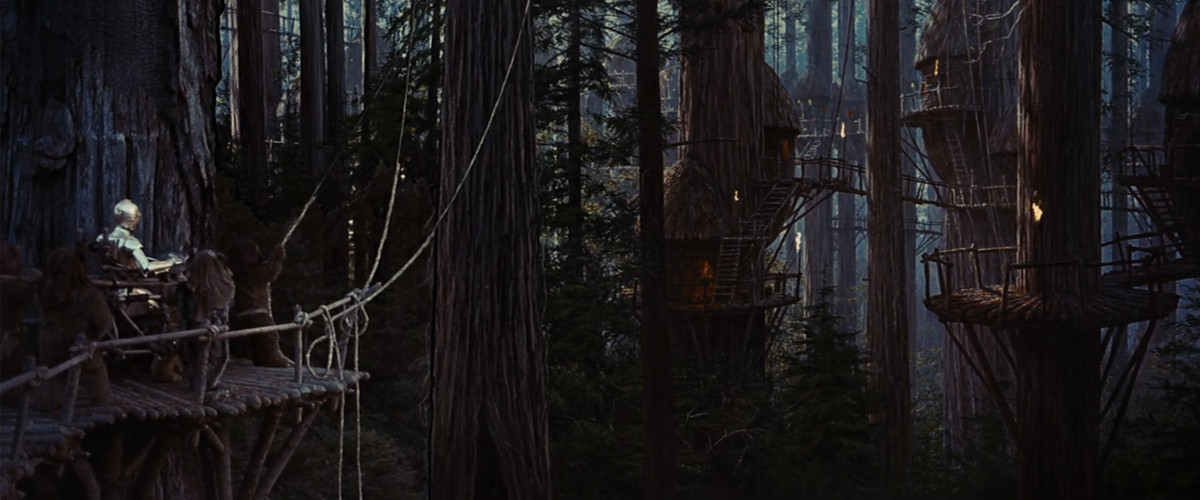 Ewoks carry C-3PO in a throne across a wooden platform in the forested Ewok villiage. 