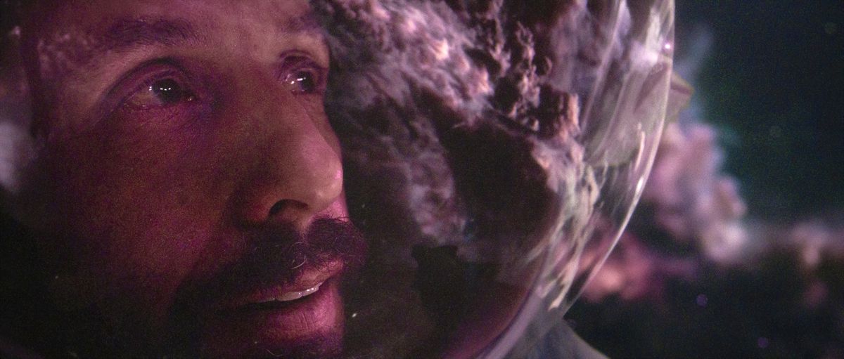 An extreme closeup on the space-suit-helmeted face of Czech astronaut Jakub (Adam Sandler), with a purple, glowing space-cloud reflected in his helmet in Netflix’s Spaceman