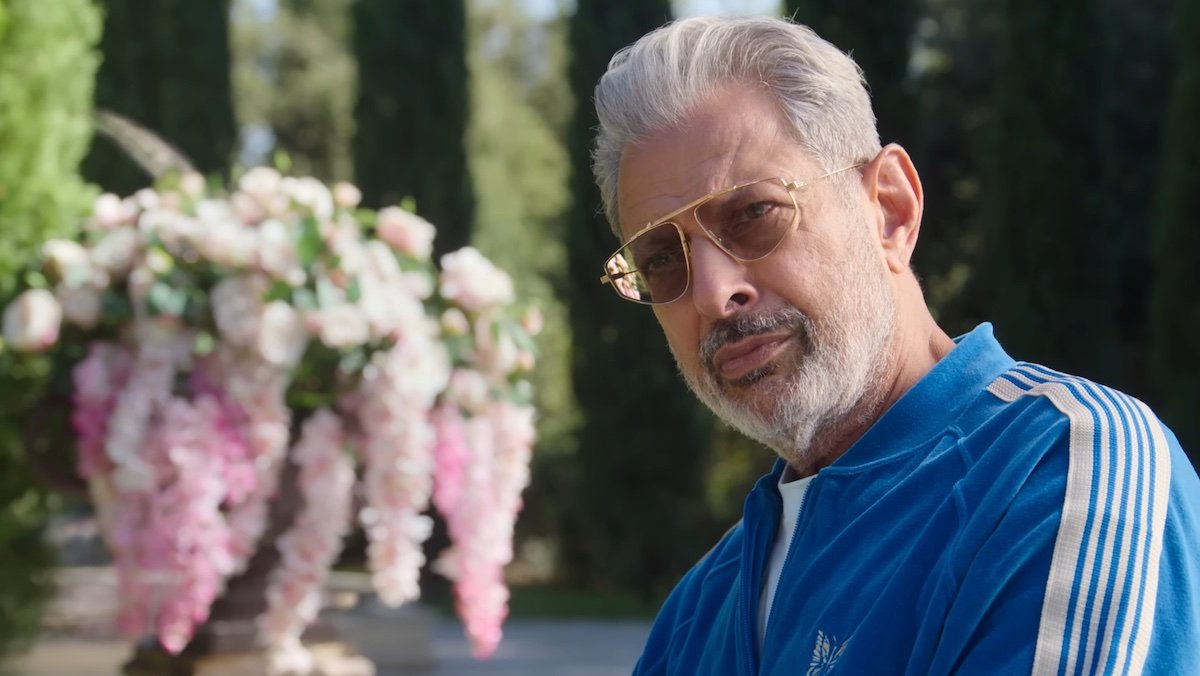Jeff Goldblum sitting outside in the sun in gold sunglasses and a blue tracksuit on KAOS