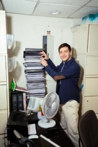 A man smiles at the camera while searching through a head-high stack of files.