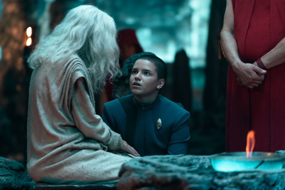 Blu del Barrio as Adira in Star Trek: Discovery. She kneels confused before a strange figure dressed in white with white hair, with red robed figures in the background. 
