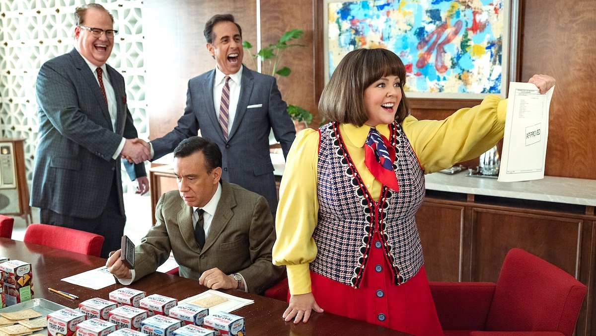 Two men shake hands while smiling as a happy woman holds up a paper and an upset an sits at a table in an office in Unfrosted the Pop-tarts movie.