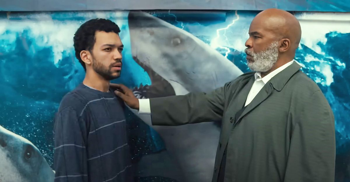 A older, bearded man puts his hand on the shoulder of a younger man in front of a shark poster.