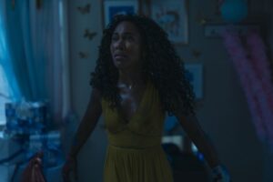 DeWanda Wise in Imaginary stands in a shadowy room wearing a yellow dress with stains on it