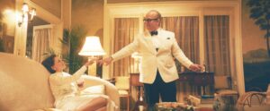 Capote, in a white dinner jacket and bow tie, stands above Babe Paley, lounging on a couch in a white dress, as he hands her a cigarette in FX’s Feud: Capote vs. The Swans.