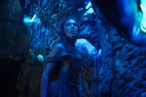 A disheveled Millie Bobby Brown stands in a cavern illuminated by a blue bioluminescent glow, looking up in awe