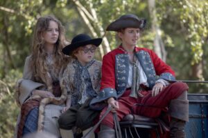 (L-R): Bo Bragason as Roxy Trotter, Florence Keen as George Trotter, and Louisa Harland as Nell Jackson sitting on the driver’s seat on a carriage in Renegade Nell.