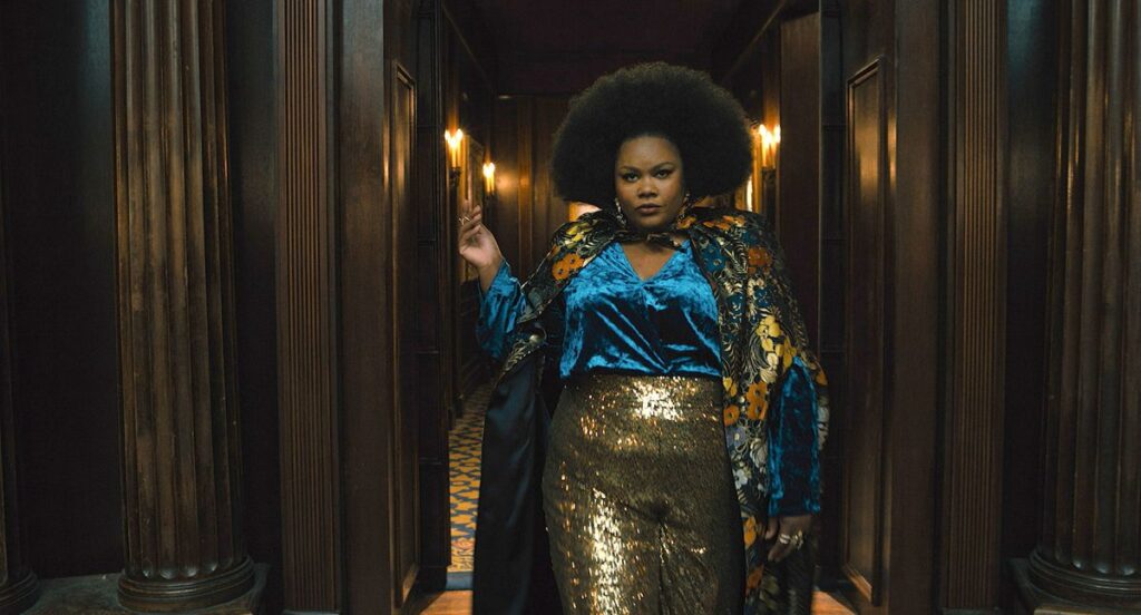 Dede (Nicole Byer), a Black woman with a large, shaped Afro, wearing gold lamé pants, an electric blue silk shirt, and a kimono-like silk flowered jacket, poses dramatically in a dark hallway in The American Society of Magical Negroes