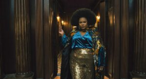 Dede (Nicole Byer), a Black woman with a large, shaped Afro, wearing gold lamé pants, an electric blue silk shirt, and a kimono-like silk flowered jacket, poses dramatically in a dark hallway in The American Society of Magical Negroes