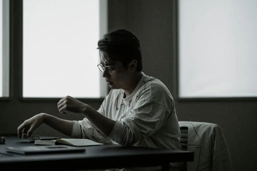 A man reads a book at a table in drab white clothing in The 8 Show