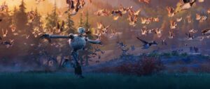 In a publicity still from DreamWorks’ animated movie The Wild Robot, a battered, bulbous white robot walks across a dark field with sunlight falling from above, holding her arms out to the sides as a flock of geese fly alongside her