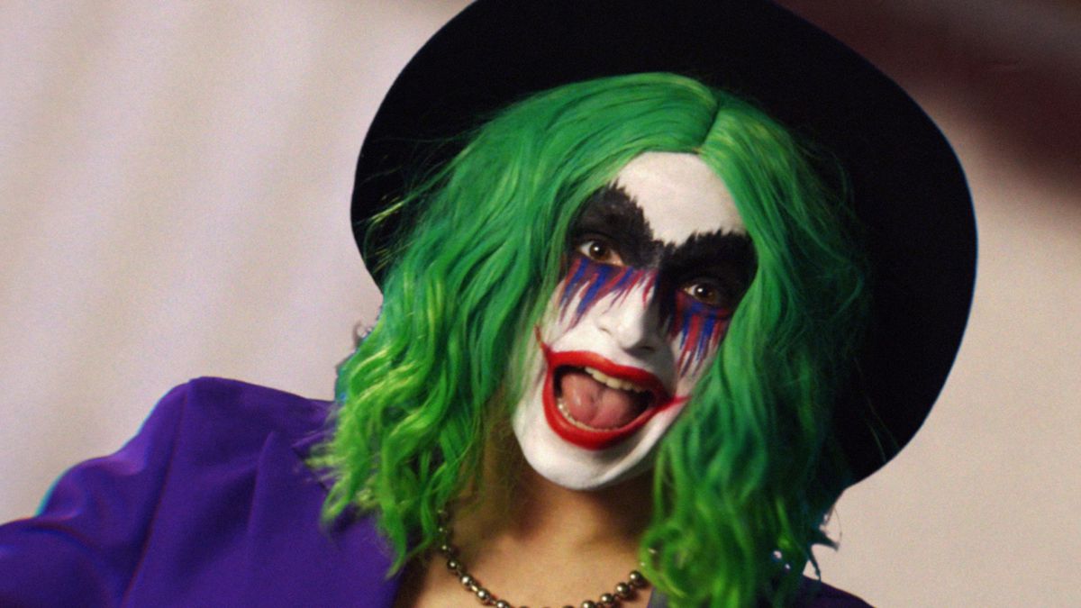 Vera Drew as Joker the Harlequin, a mashup of Joker and Harley Quinn, in green wig, clown face paint with a big red lipstick smile, a purple suit jacket, and black hat in The People’s Joker