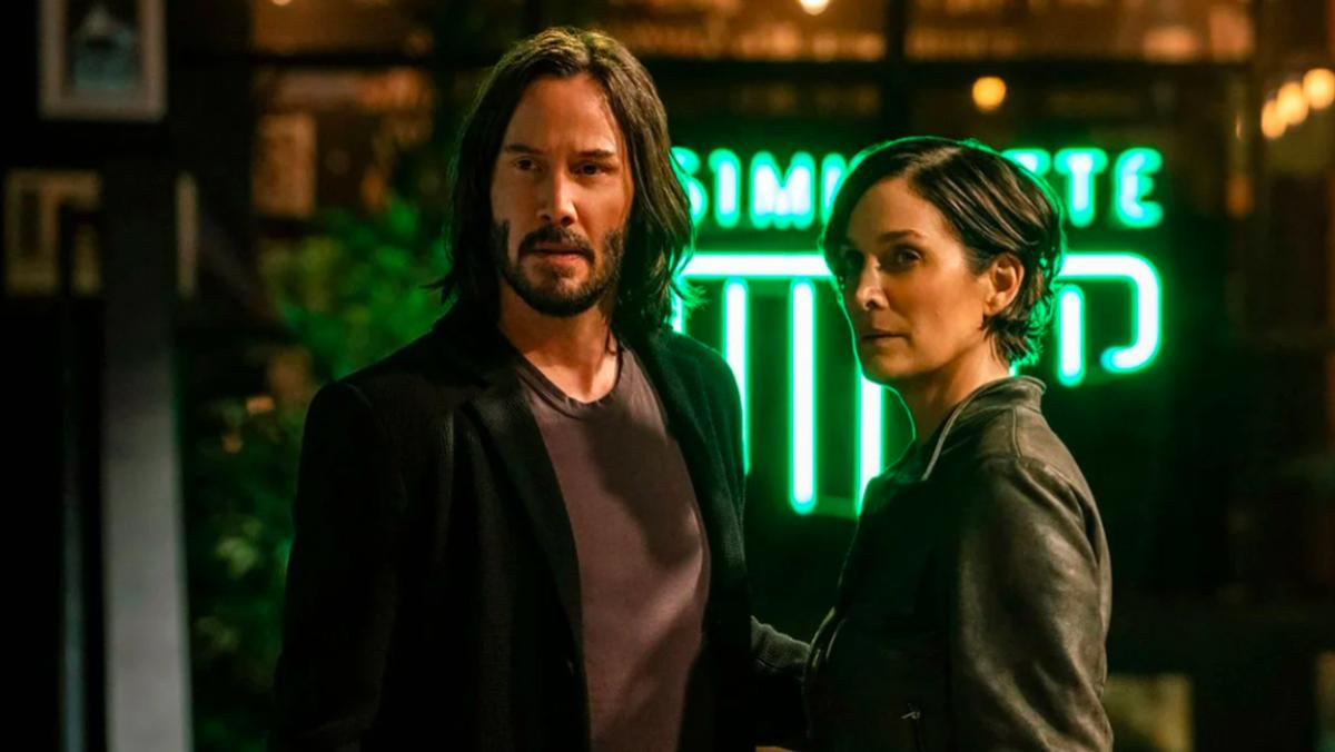 A still from The Matrix Resurrections shows Keanu Reeves as Neo and Carrie Anne Moss as Trinity standing in front of a coffee shop sign that says Simulatte