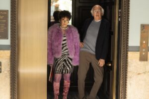 A woman in a fuzzy purple coat stands next to a man in a doorway.
