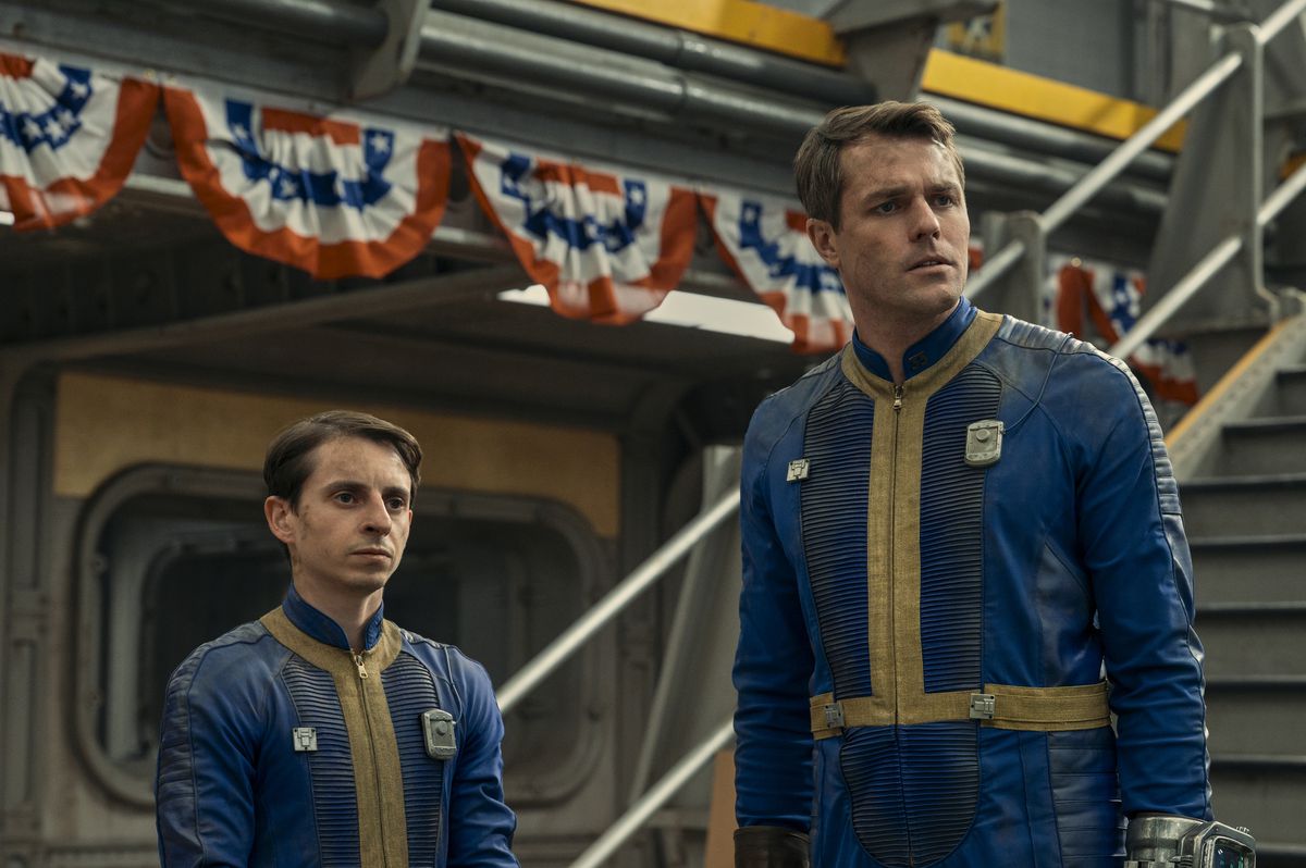 Two men in a blue and yellow uniforms standing side by side in front of a staircase with red, white, and blue decorations beside it in the Fallout TV series.