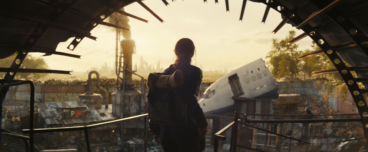 A young woman with a backpack staring out at a town made of scavenged metal in the Fallout TV series.