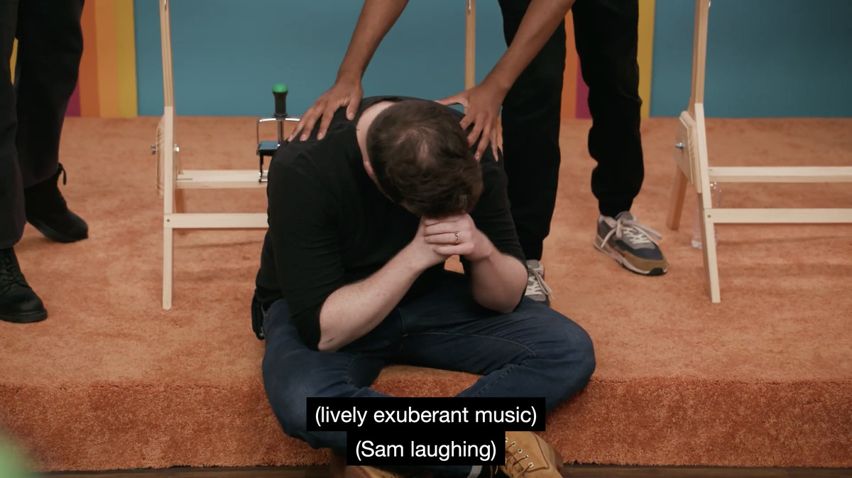 A screencap of Game Changer’s “Bingo,” in which Brennan Lee Mulligan sits on the floor with his head in his hands. Closed captioning reads “(lively exuberant music)” and “(Sam laughing).”