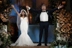 A woman in a wedding dress stands next to a man in a black tuxedo, both holding their arms up triumphantly.