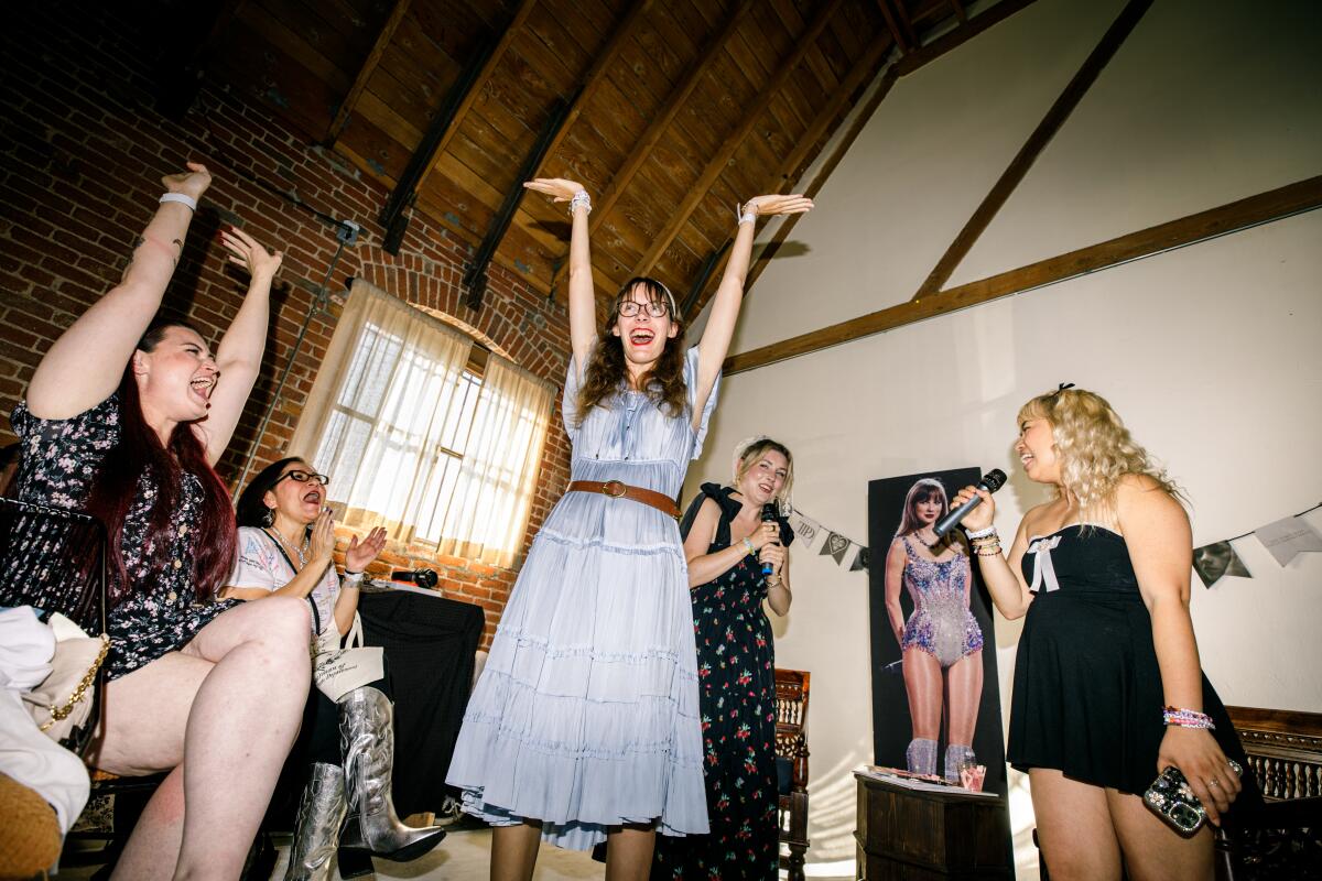 A photo of a woman posing and smiling and others cheering around her at the Taylor Swift listening party