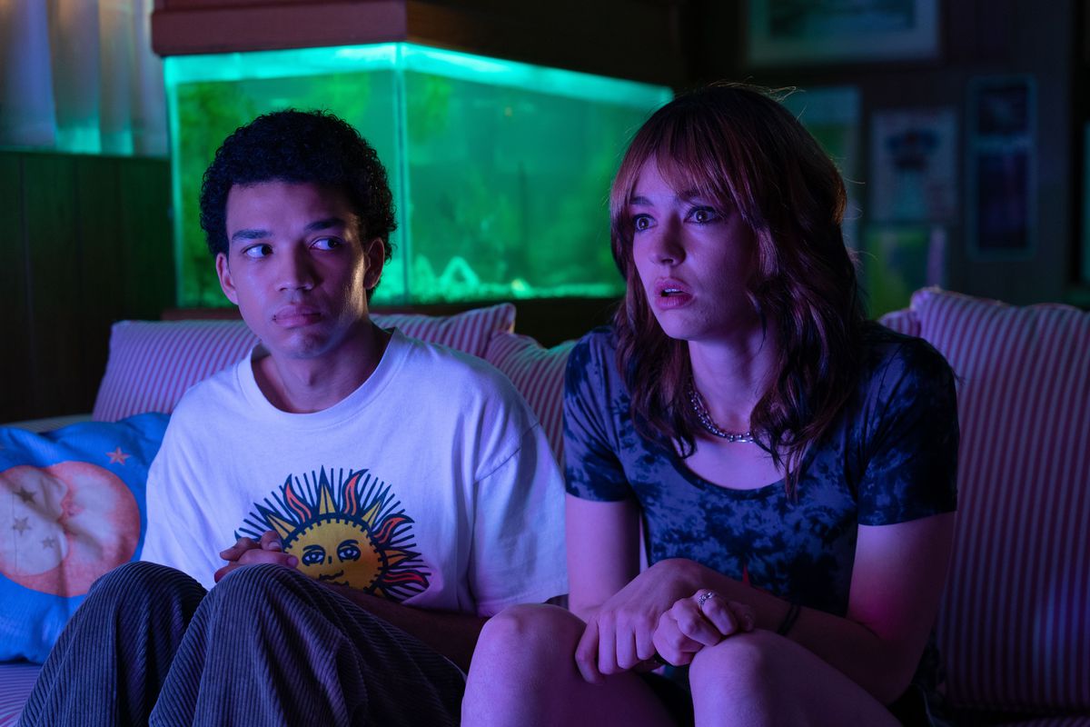 Justice Smith and Bridgette Lundy-Paine sit in a neon-light drenched room looking toward a bright screen in I Saw the TV Glow