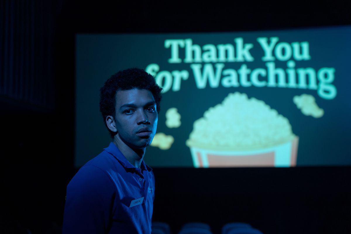 Justice Smith stands in front of a movie theater screen that says “Thank You for Watching” with a big ol’ bucket of popcorn in I Saw the TV Glow