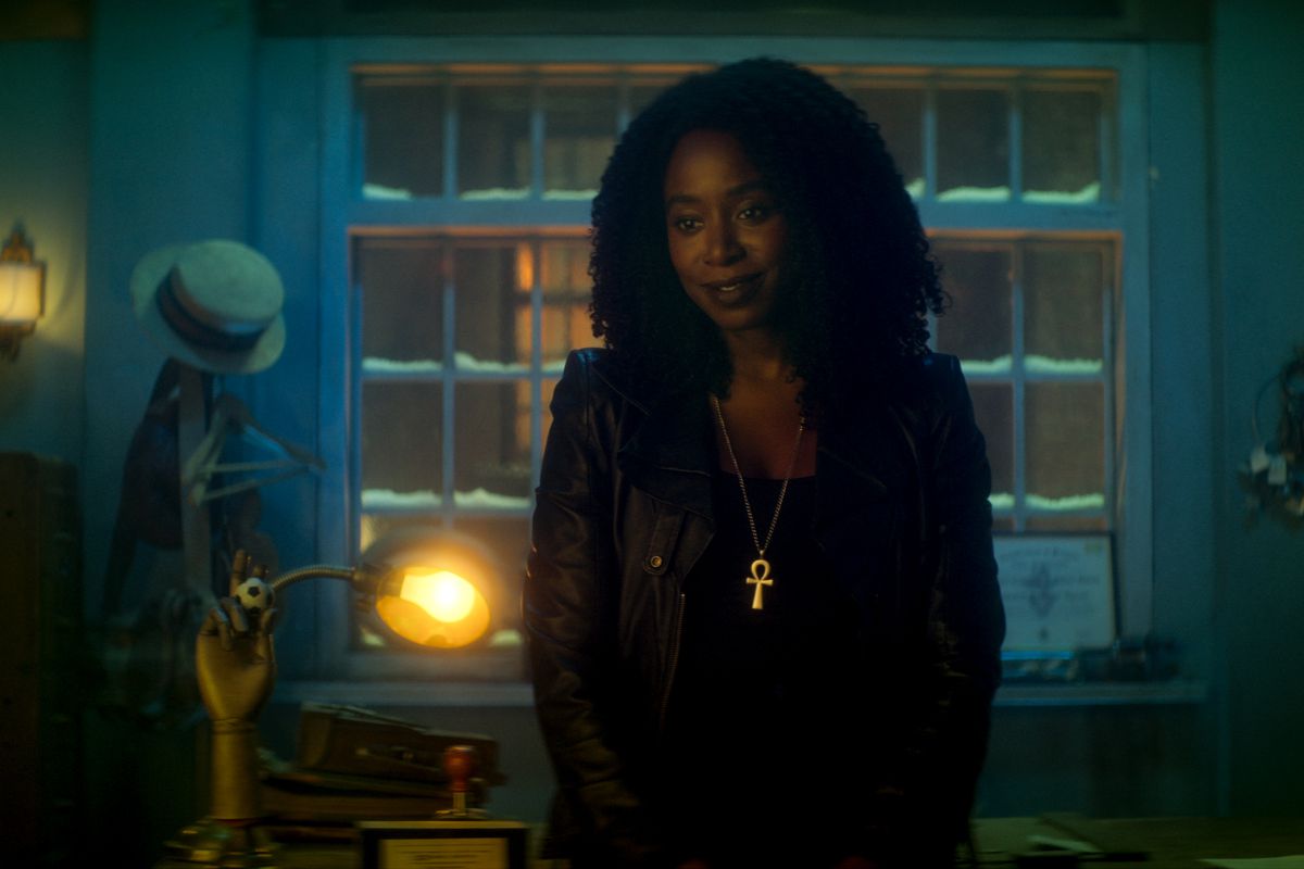 Kirby Howell-Baptiste as Death in Dead Boy Detectives. She’s dressed in all black, with a gold ankh necklace, standing in a blue-tinged room.