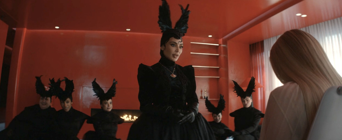 Kim Kardashian in a garish black feathered headdress leering over Emma Roberts. Behind her is a gaggle of women in the same headdress. They all stand in a bright red modern loft. 