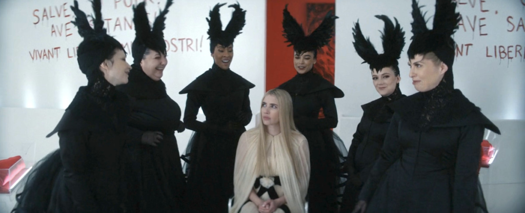 Emma Roberts sits in a wheelchair, surrounded by six women in black feathered headdresses