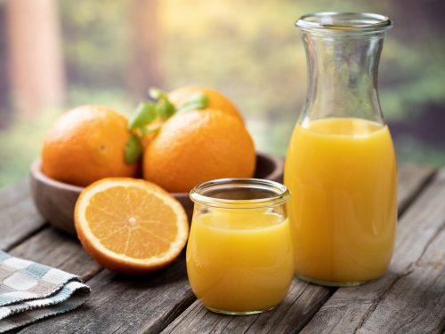 An orange juice glass and carafe set next to a sliced orange and bowl of whole oranges