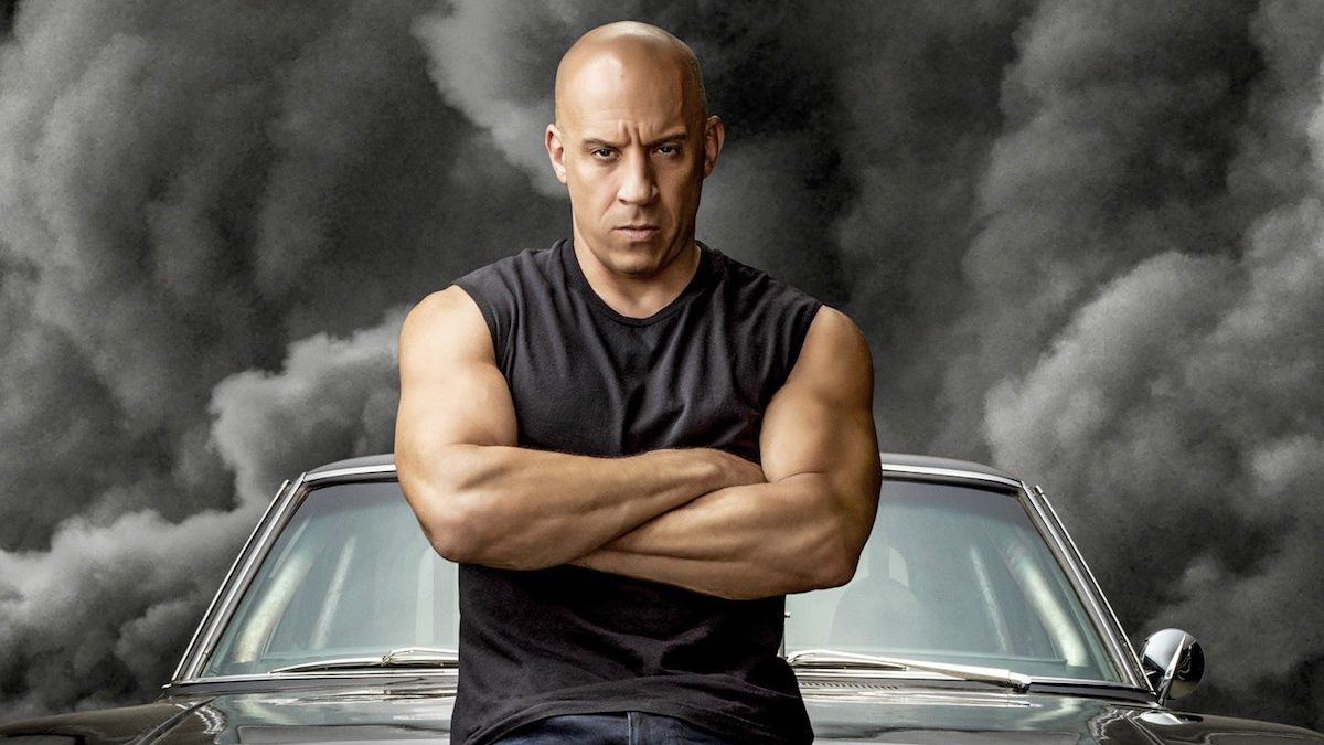 Vin Diesel in a black sleeveless t-shirt standing against a black car with black smoke behind it