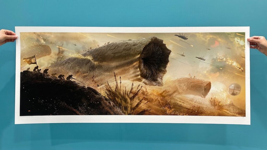 Two hands near a blue wall hold up a giant Dune: Part Two poster showing a Fremen sandworm attack