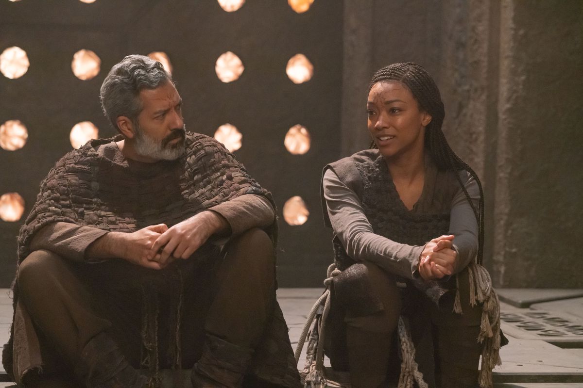 L-R Alfredo Narciso as Ohvahz and Sonequa Martin-Green as Michael Burnham in Star Trek: Discovery. They are wearing hand-made alien garments, and conversing calmly while sitting on the floor in a stone room. 