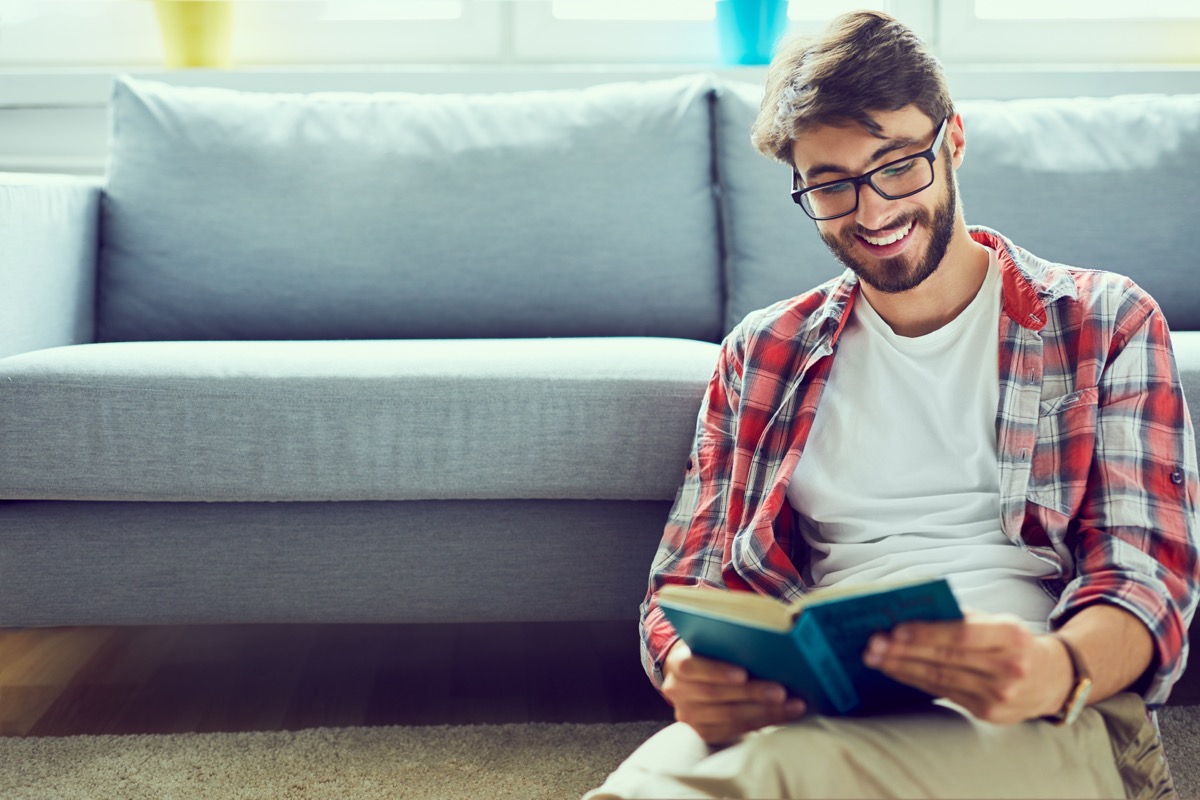 Man sitting on floor against couch reading and smiling at book