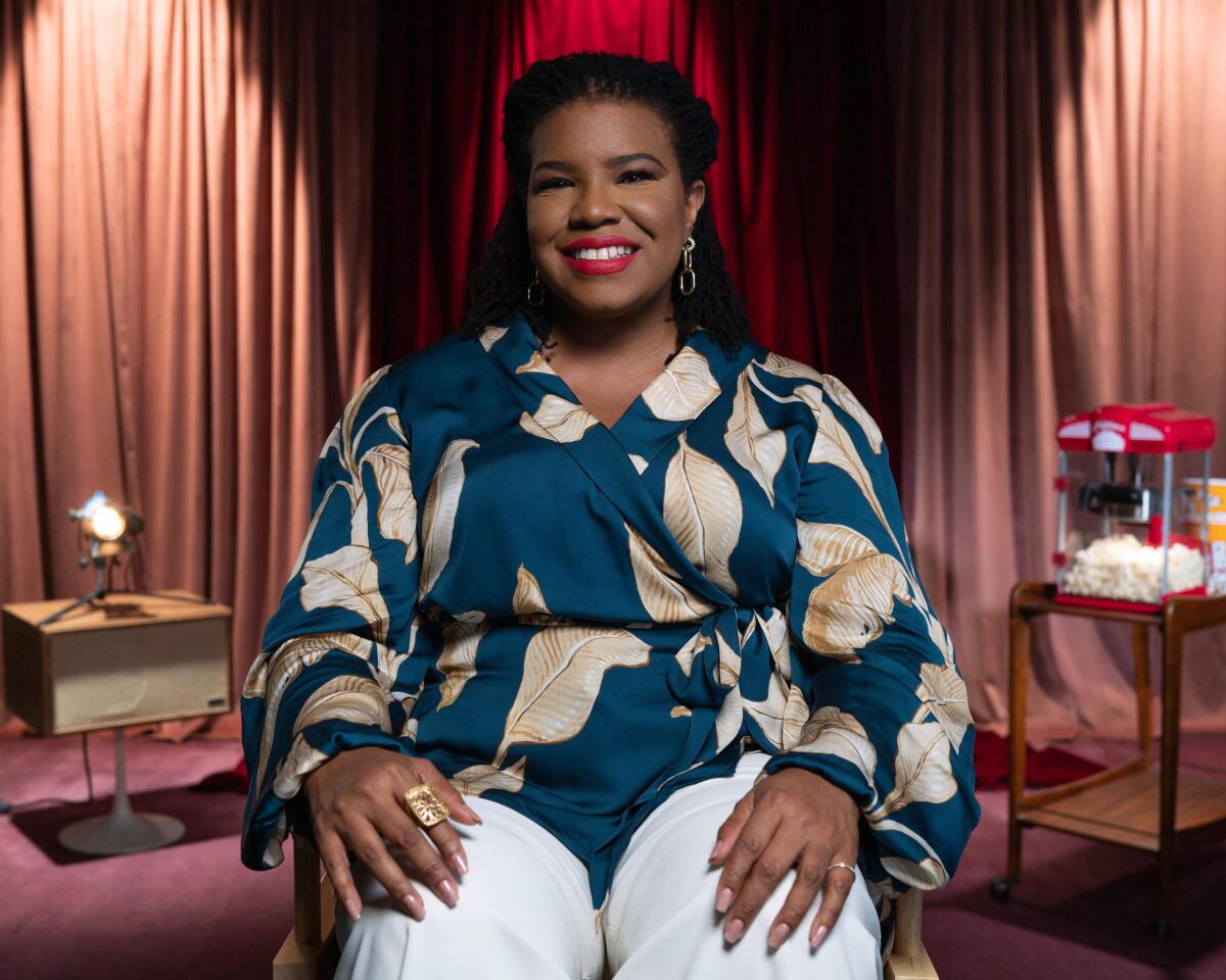 A woman in a blue floral top and white pants sitting with her hands on her knees.