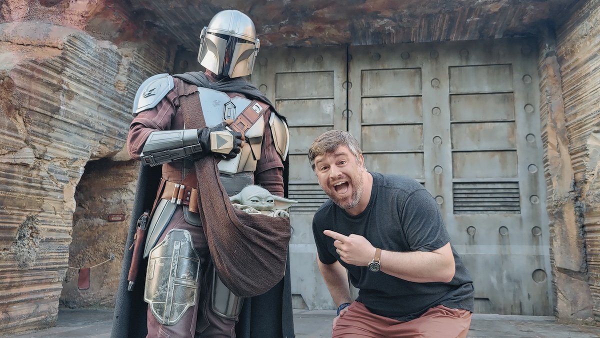 A happy man kneels down pointing at Grogu in The Mandalorian's sack at Galaxy's Edge
