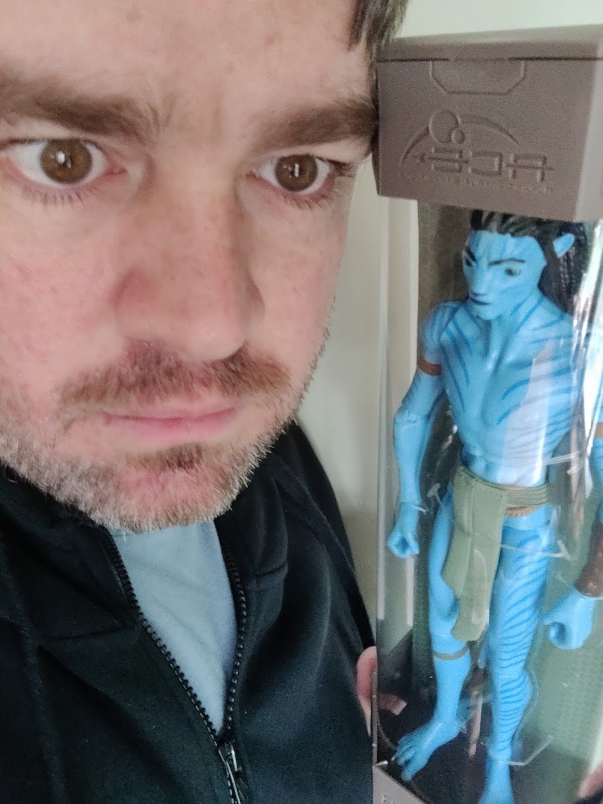 A bearded man with a crazed look holds up a blue Avatar Na'vi action figure