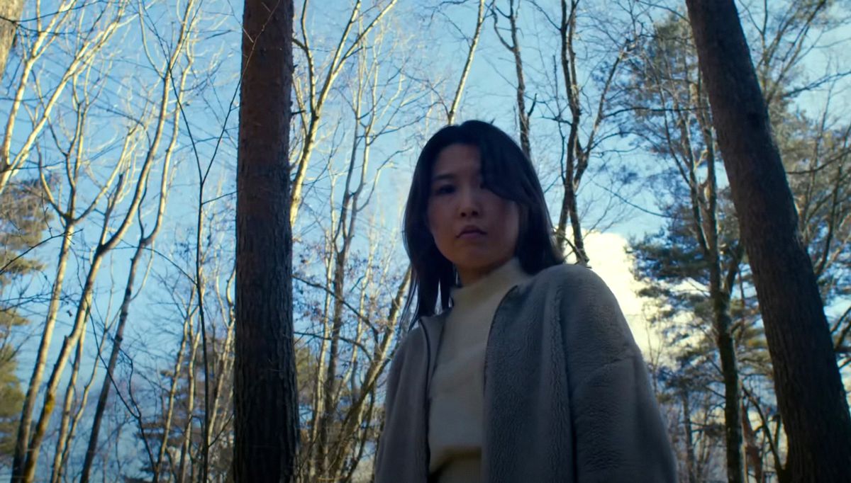 Mayuzumi (Ayaka Shibutani), a Japanese woman in a white shirt and grey cardigan, stands in the woods, looking downward at the camera, in Ryûsuke Hamaguchi’s Evil Does Not Exist