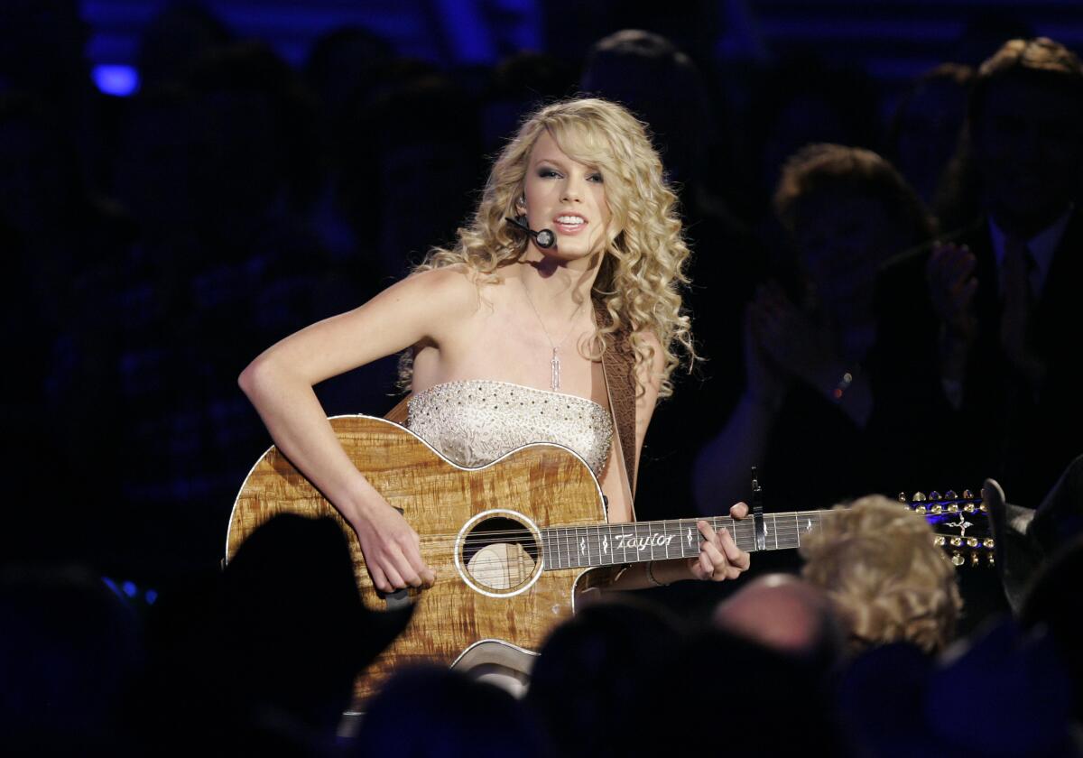 Taylor Swift sitting and playing an acoustic guitar on a darkened stage with long curly hair and clad in a strapless top