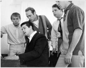 Four men singing behind a fifth man seated and playing the piano