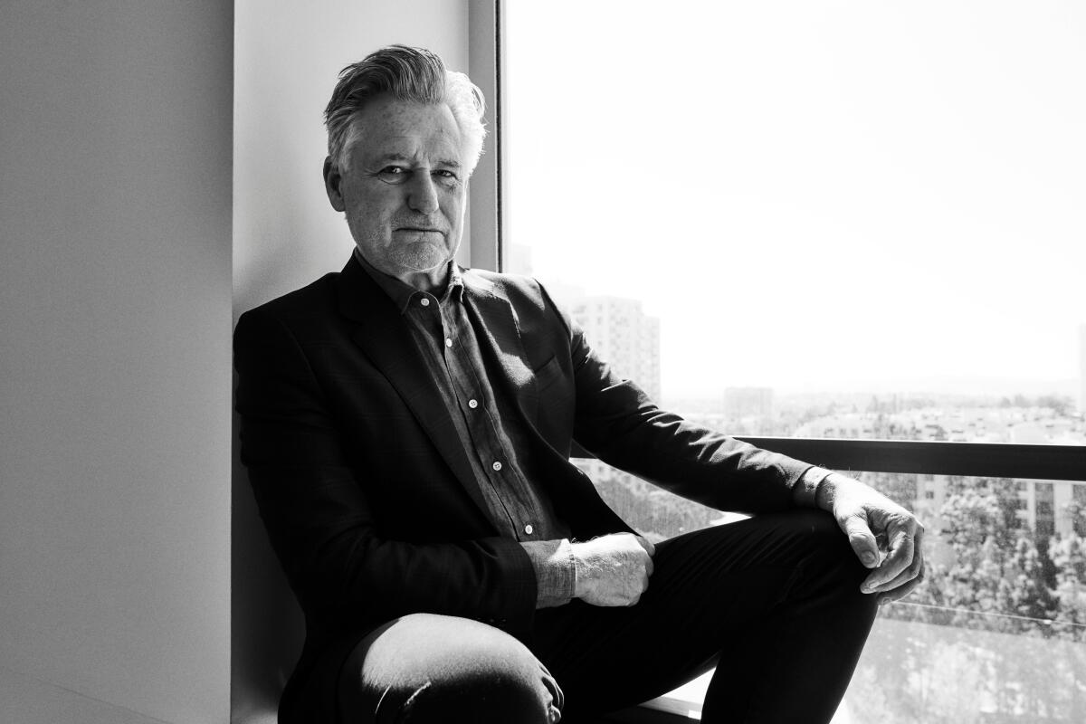 Bill Pullman sits on a chair on a balcony for a black-and-white portrait.