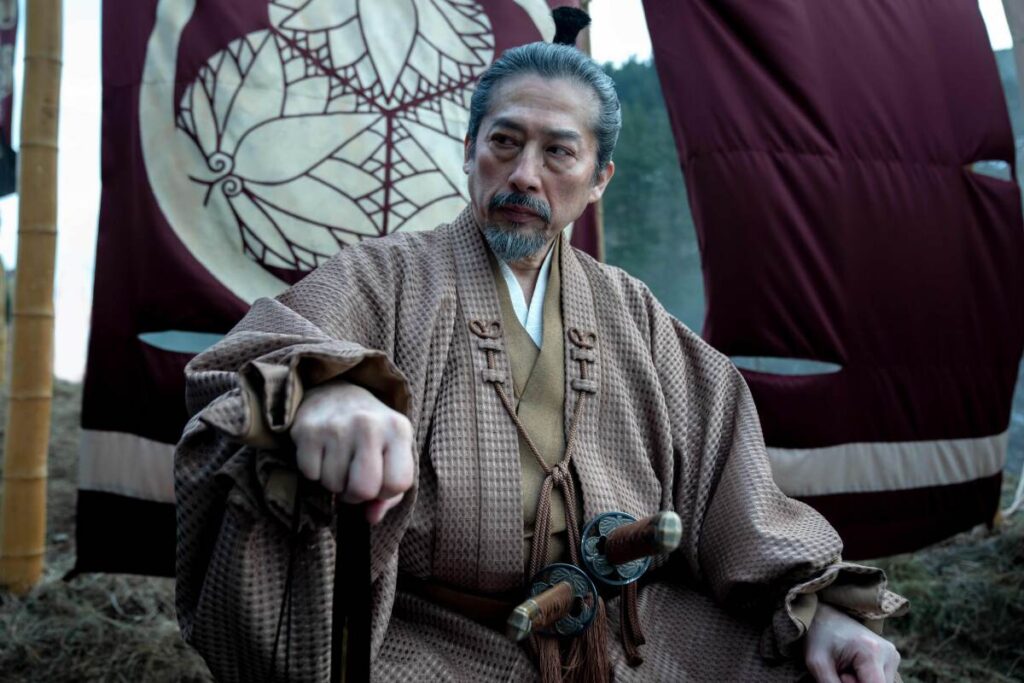 Hiroyuki Sanada as Yoshii Toranaga sitting with his fist out in front of a flag bearing a family crest in “Shōgun."