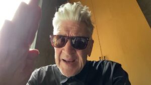 David Lynch, wearing sunglasses, gives a salute-wave to the camera.