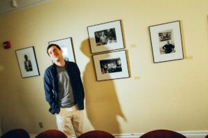 Stand-up comedian Alex Edelman stands in front of framed black-and-white photographs for a portrait.