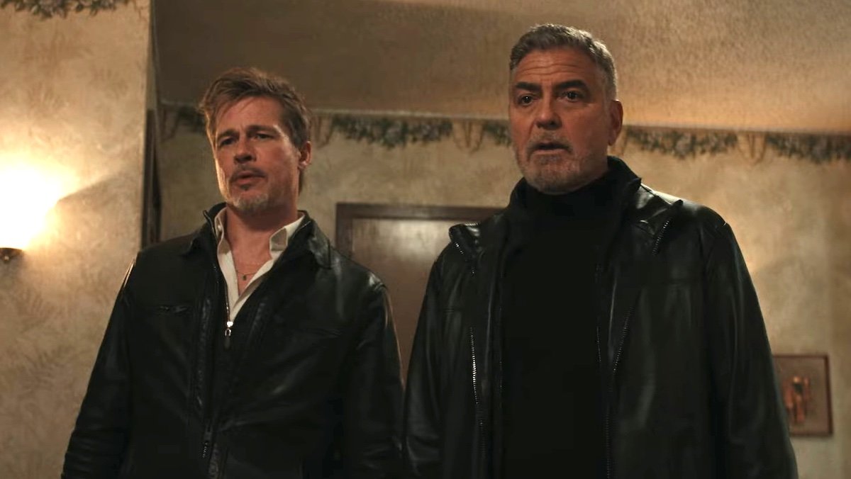 Brad Pitt and George Clooney both dressed in black inside a hotel room in Wolfs