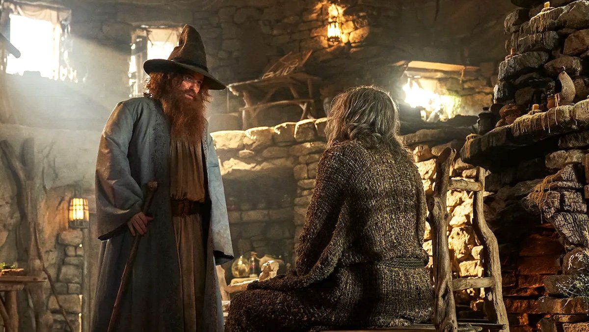 Tom Bombadil with his long hair, beard, pointed hat, robe, and cane speaks to a sitting Stranger inside a cottage on The Rings of Power