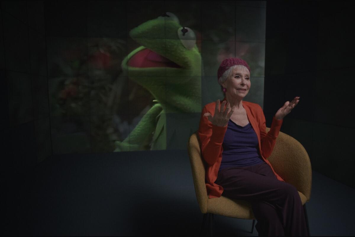 An image of Kermit the frog on a big screen behind Rita Moreno sitting in a chair gesticulating.